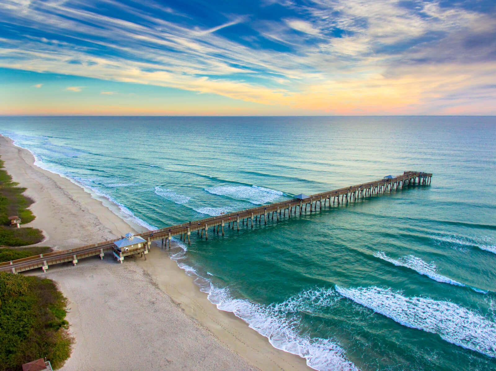 A serene beach scene with a long wooden pier stretching into the turquoise ocean, framed by a vibrant sunset sky with wispy clouds