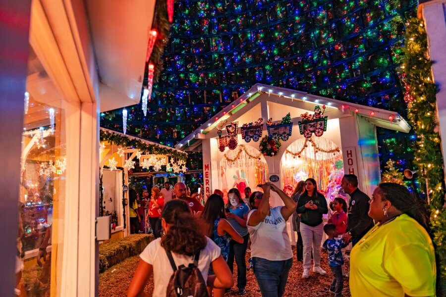 A festive crowd gathers around a brightly lit Christmas village, adorned with colorful lights and decorations, creating a magical holiday atmosphere