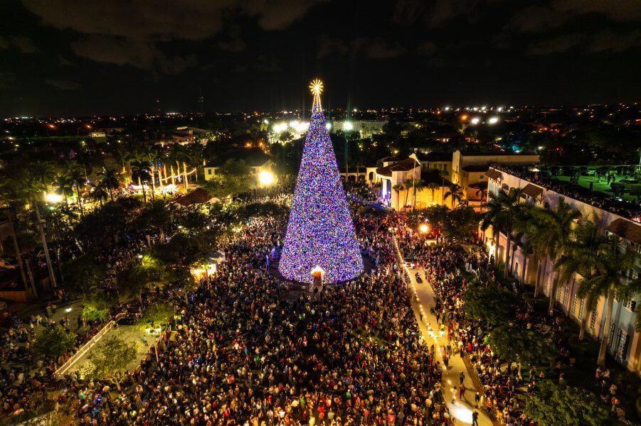 A massive, brightly lit Christmas tree stands tall in the center of a bustling city square, surrounded by a sea of people enjoying the festive atmosphere