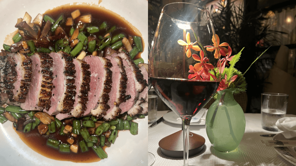 A beautifully plated dish of seared duck breast with green beans and mushrooms in a rich sauce, accompanied by a glass of red wine and a vibrant flower arrangement
