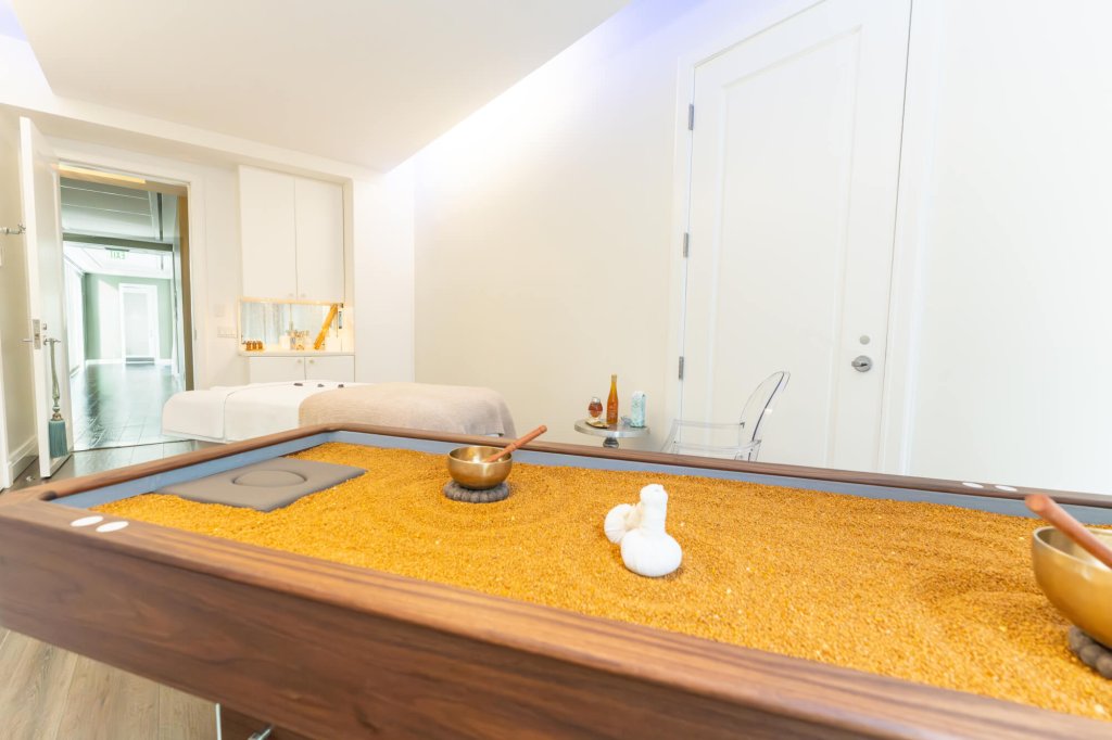 Une salle de spa sereine avec une table en bois recouverte de sable doré, avec un bol, des cuillères et des pierres blanches, ainsi qu'une table de massage à l'arrière-plan.
