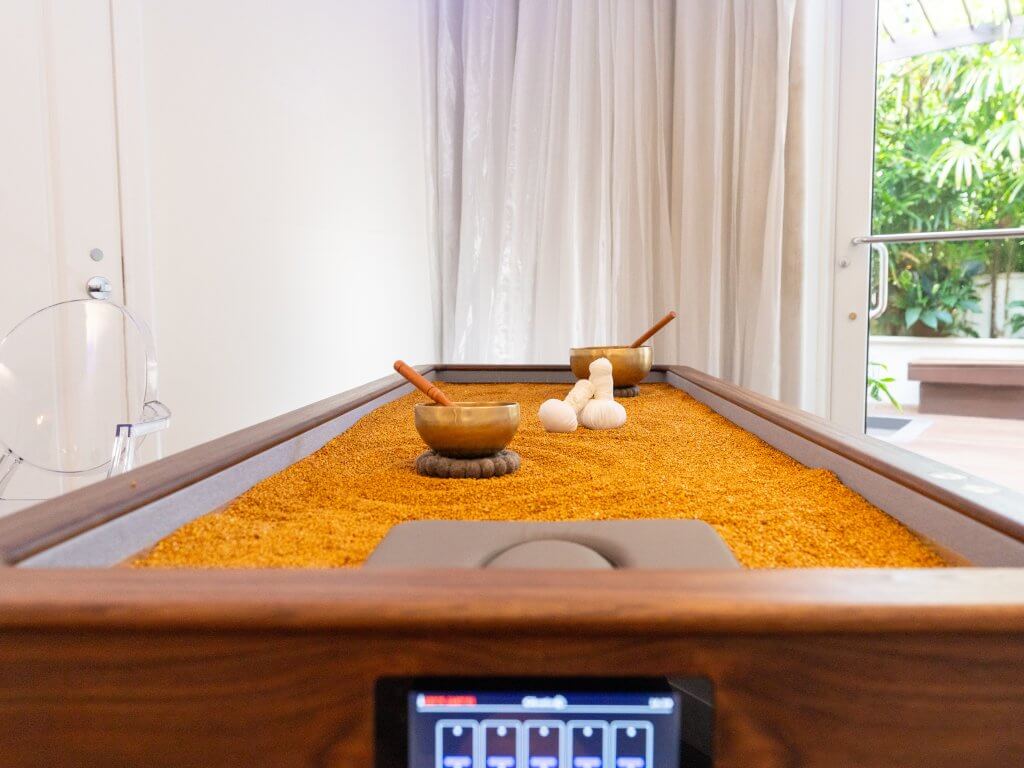 A wooden table with a sand surface, featuring bowls, a digital display, and a clear chair, set against a backdrop of white curtains and a glass door leading to a garden