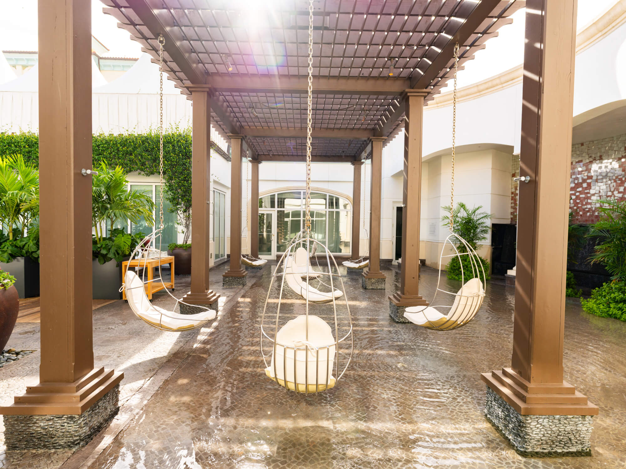 A serene outdoor lounge area with hanging egg chairs suspended from a wooden pergola, surrounded by lush greenery and a reflective water feature