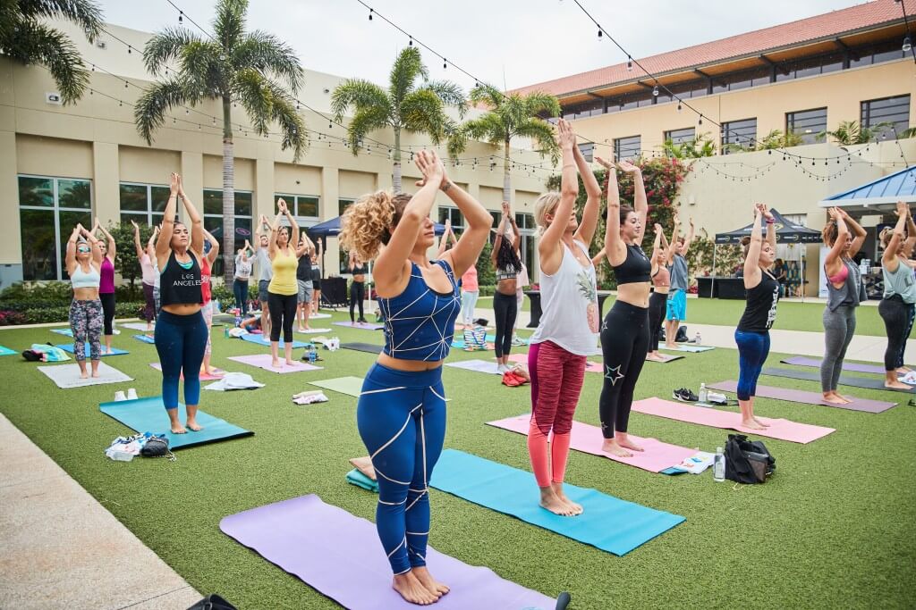 A group of women practicing yoga outdoors on colorful mats in a grassy area with palm trees and a building in the background
