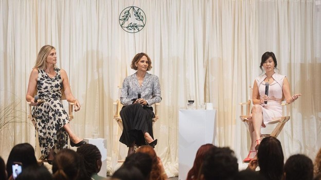 Three women in elegant attire sit on stage chairs, engaged in a panel discussion before an audience