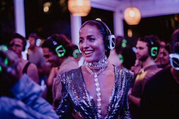A woman in a sparkling silver dress and headphones smiles brightly at a crowded, dimly lit party