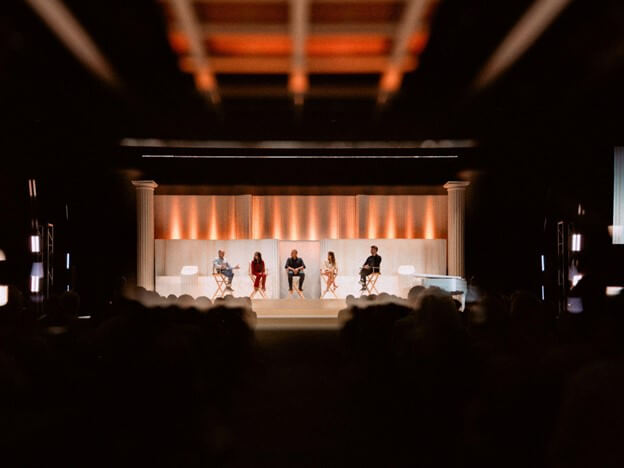 Four individuals are seated on a stage with a minimalist backdrop, addressing an audience in a dimly lit auditorium