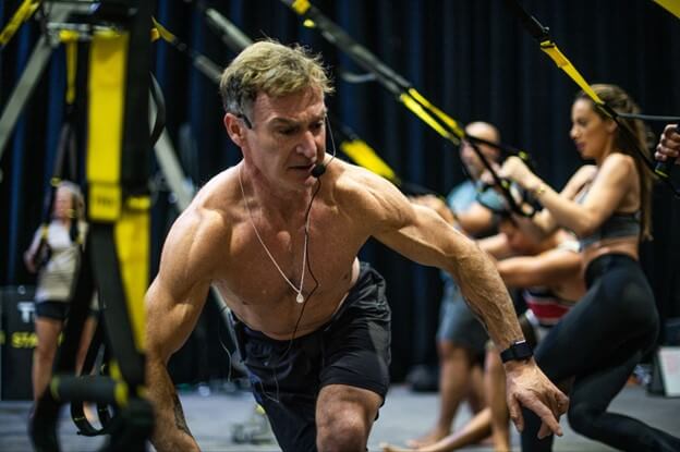 A shirtless man with a microphone headset leads an intense workout class using suspension training equipment