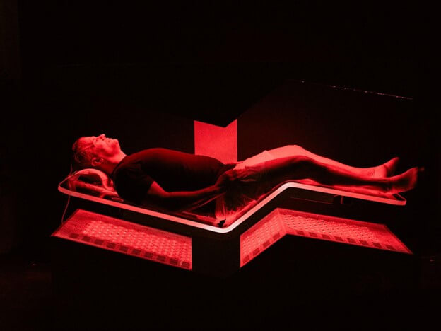 A person is lying on a futuristic bed illuminated by red light, appearing to be in a state of relaxation or medical treatment