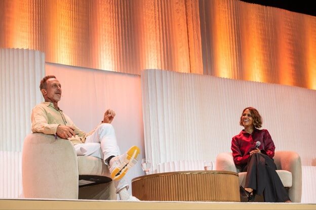 Two individuals are seated on stage, engaged in conversation, with a modern backdrop of curved white panels and warm orange lighting