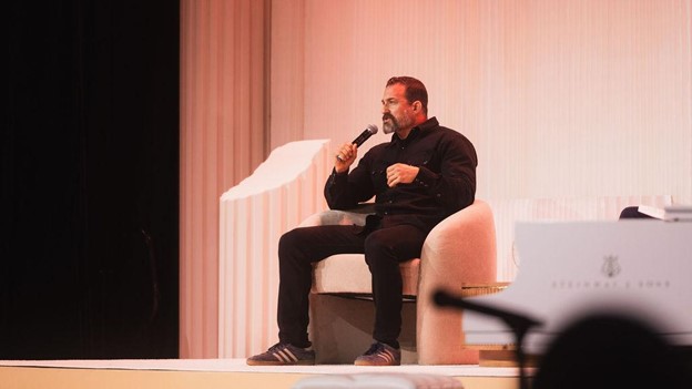 A man in a black suit sits on a white chair, holding a microphone and addressing an audience
