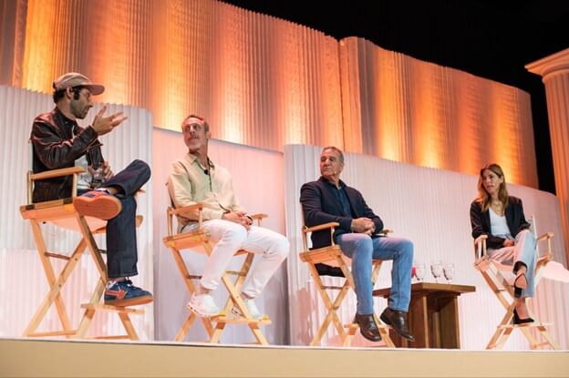 Four individuals are seated on director's chairs on a stage with a backdrop of warm, ambient lighting