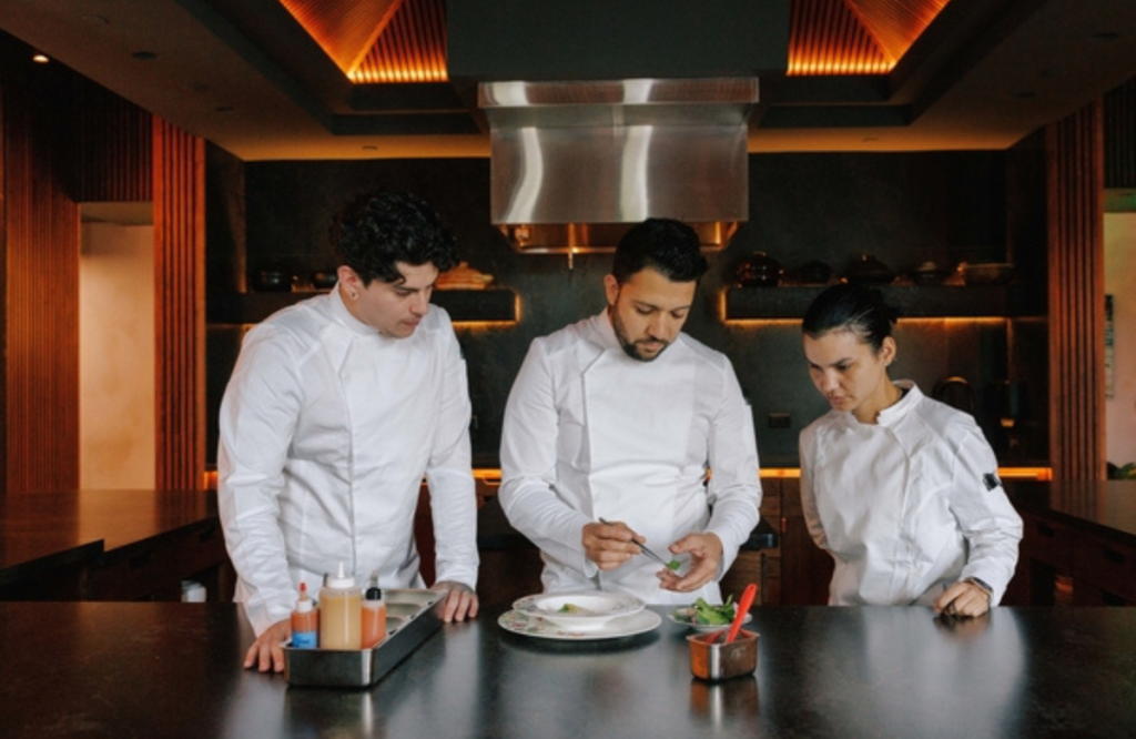 Three chefs in white uniforms working together on a dish in a modern kitchen