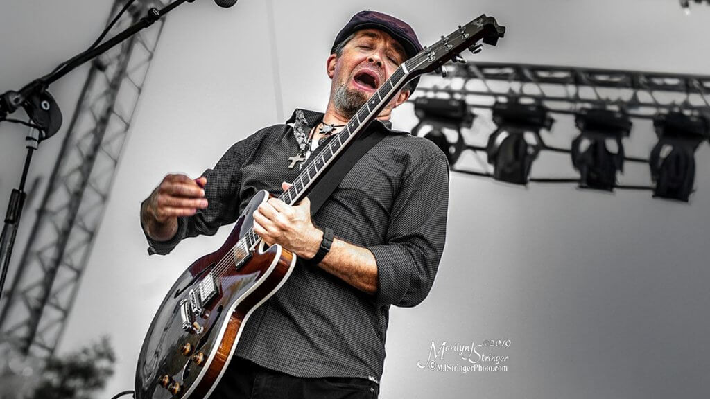 A musician passionately plays an electric guitar on stage, wearing a black hat and a cross necklace