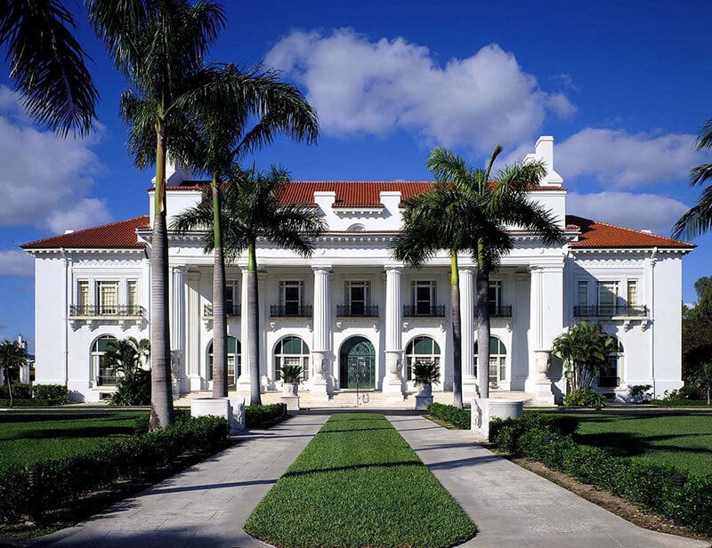 A grand white mansion with columns and a red-tiled roof, surrounded by palm trees and lush greenery