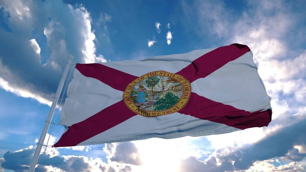 The flag of Florida, featuring a white cross with red and white stripes, flutters against a backdrop of a partly cloudy blue sky