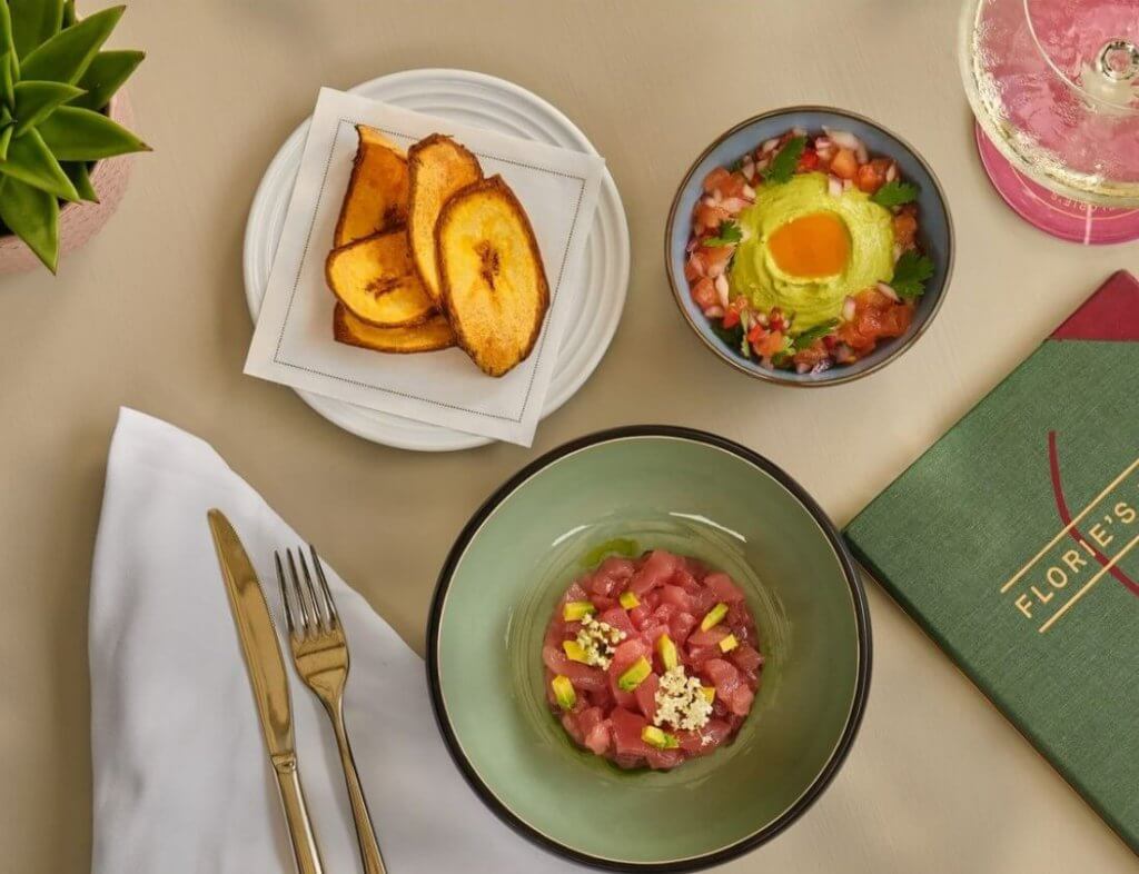 Una mesa puesta con una variedad de platos coloridos, entre los que se incluyen chips de plátano, un tartar de atún y una ensalada de vivos colores, acompañada de una carta de menú verde y una copa de vino espumoso