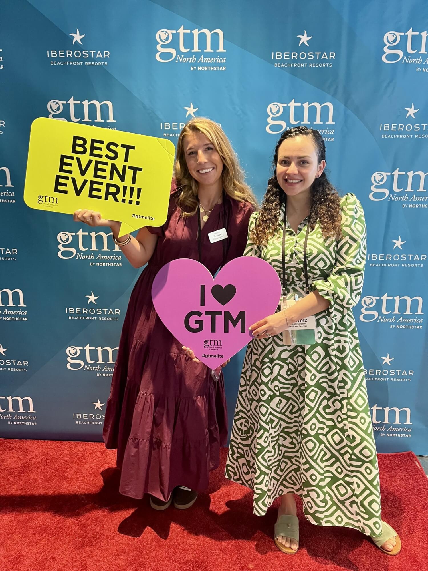 Two women holding event signs in front of a branded step and repeat backdrop