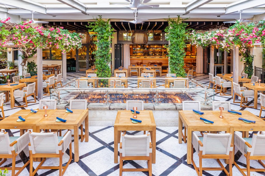 Um restaurante ao ar livre com mesas de madeira, cadeiras brancas e uma fogueira central cercada por uma vegetação exuberante e flores cor-de-rosa