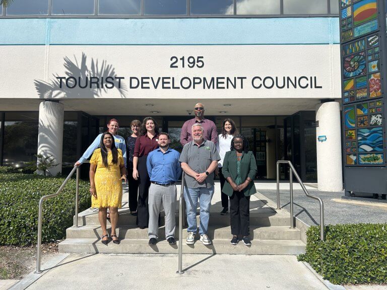 Un groupe diversifié de dix personnes se tient devant le bâtiment du Conseil du développement touristique, souriant et posant pour une photo de groupe.