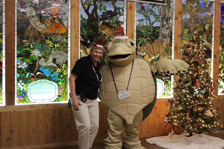 A woman in a festive hat stands next to a person in a turtle mascot costume, both smiling in front of a colorful stained glass window and a decorated Christmas tree