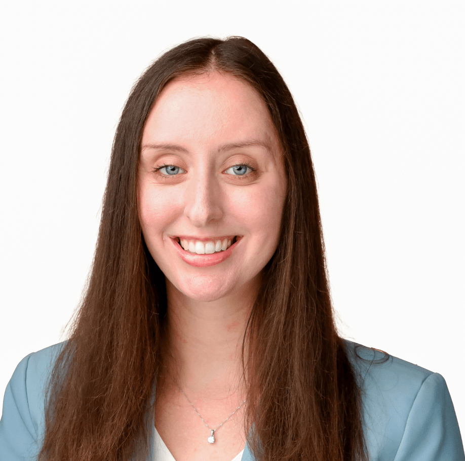 A woman with long brown hair and blue eyes smiles warmly at the camera, wearing a light blue blazer and a delicate necklace