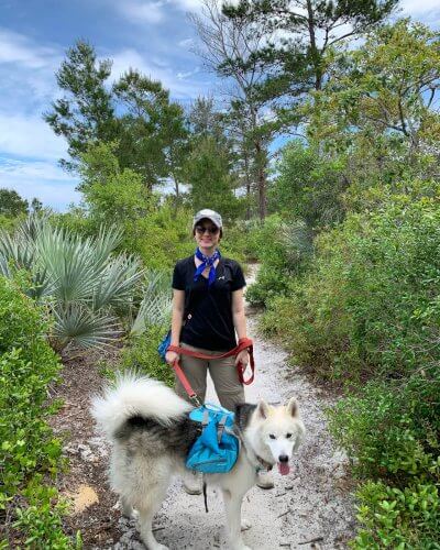 Eine Frau und ihr Hund wandern auf einem Pfad durch einen üppigen Wald, wobei die Frau ein blaues Halstuch trägt und der Hund einen blauen Rucksack mit sich führt.