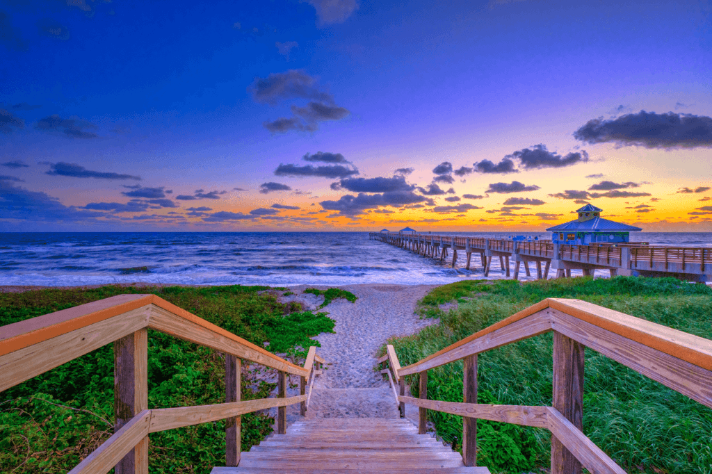 Una escalera de madera desciende hasta una tranquila playa al atardecer, con un largo muelle que se adentra en el océano y un pequeño mirador en su extremo