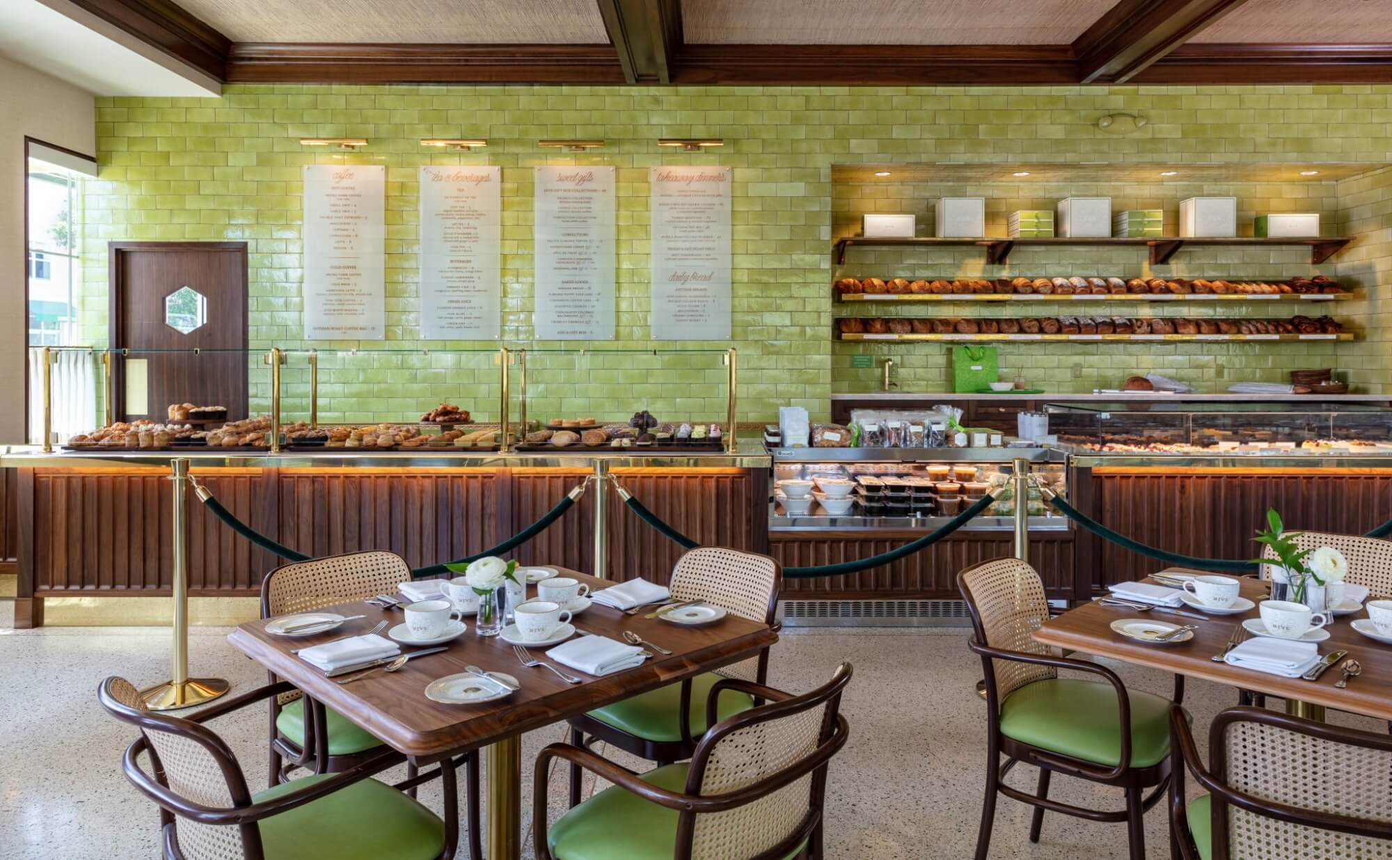 A cozy bakery cafe with a green tiled wall, wooden tables and chairs, and a display case filled with pastries and bread