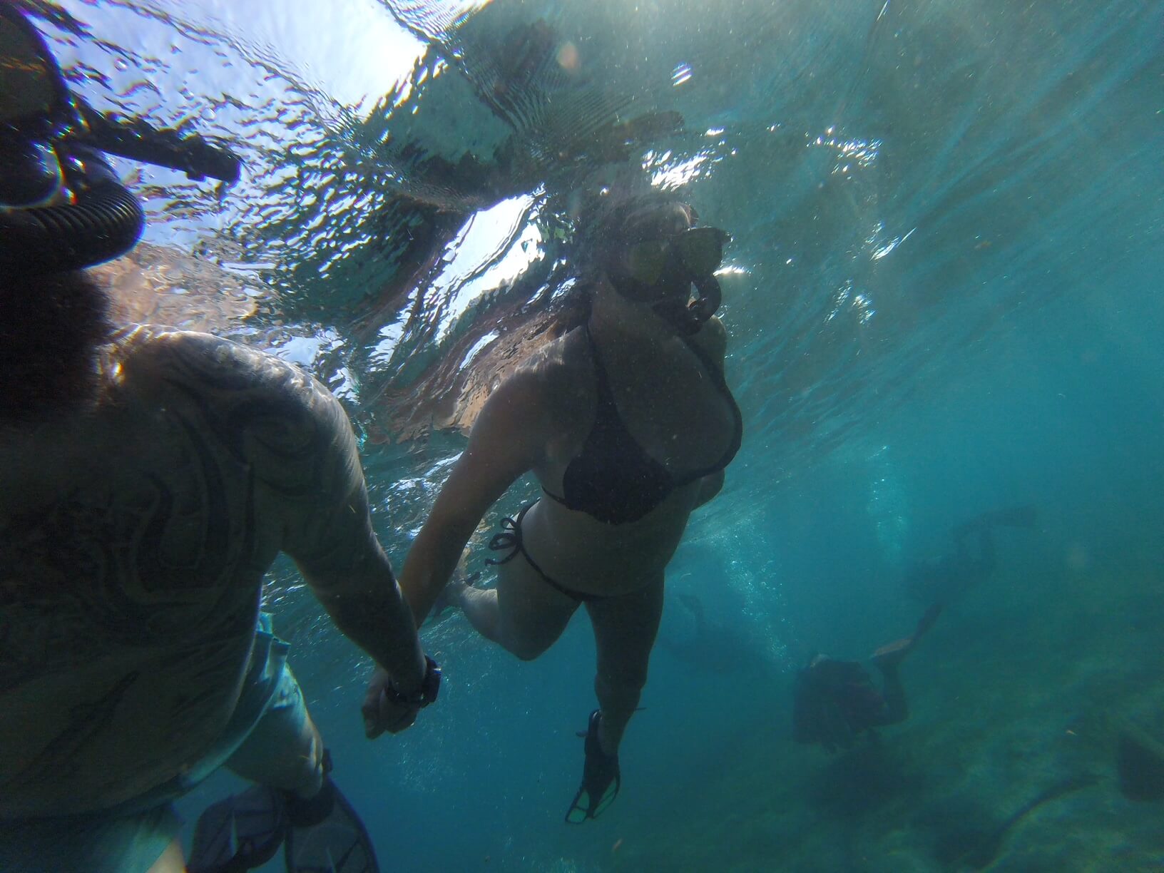 A couple swims underwater, holding hands, with sunlight filtering through the water's surface above them