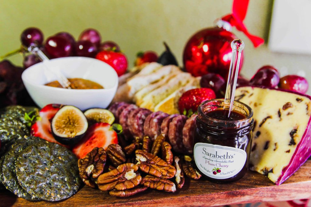 Uma tábua de charcutaria festiva com uma variedade de queijos, carnes curadas, frutas, nozes, biscoitos e um pote de geleia de ameixa e cereja
