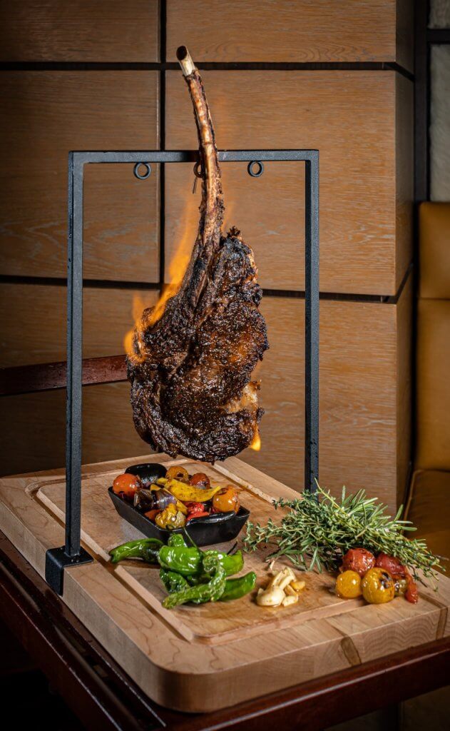 A perfectly cooked tomahawk steak with a flaming bone, served on a wooden board with a variety of roasted vegetables and fresh herbs