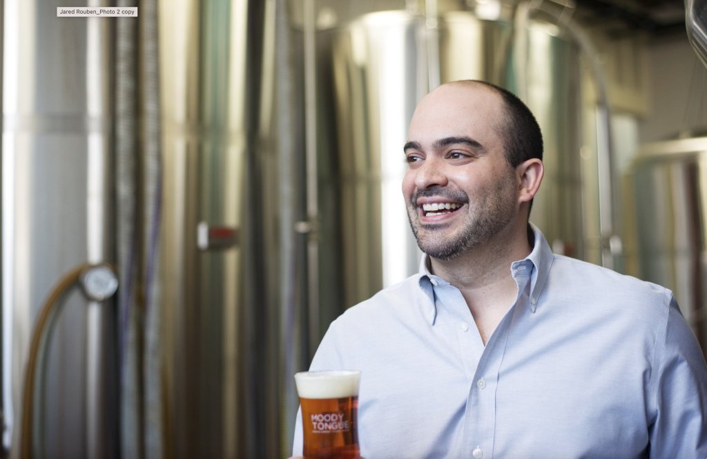A man in a brewery holding a glass of beer with the label 'Moody Tongue'