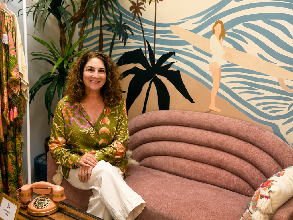 A woman sits on a pink couch in a room with a tropical mural, wearing a colorful floral shirt and white pants