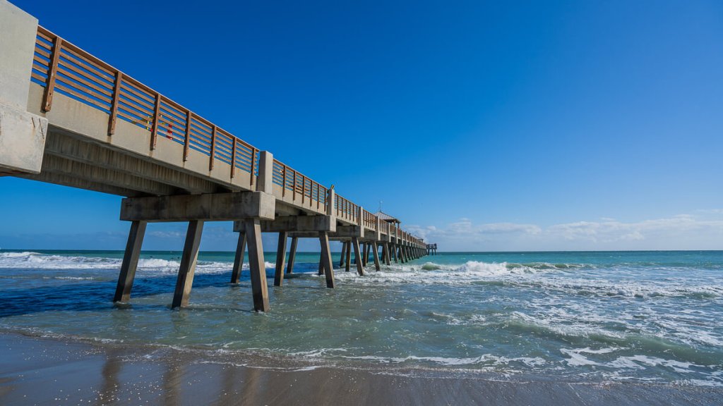 Un largo muelle de madera se adentra en el océano, con las olas rompiendo contra sus pilares bajo un cielo azul y despejado