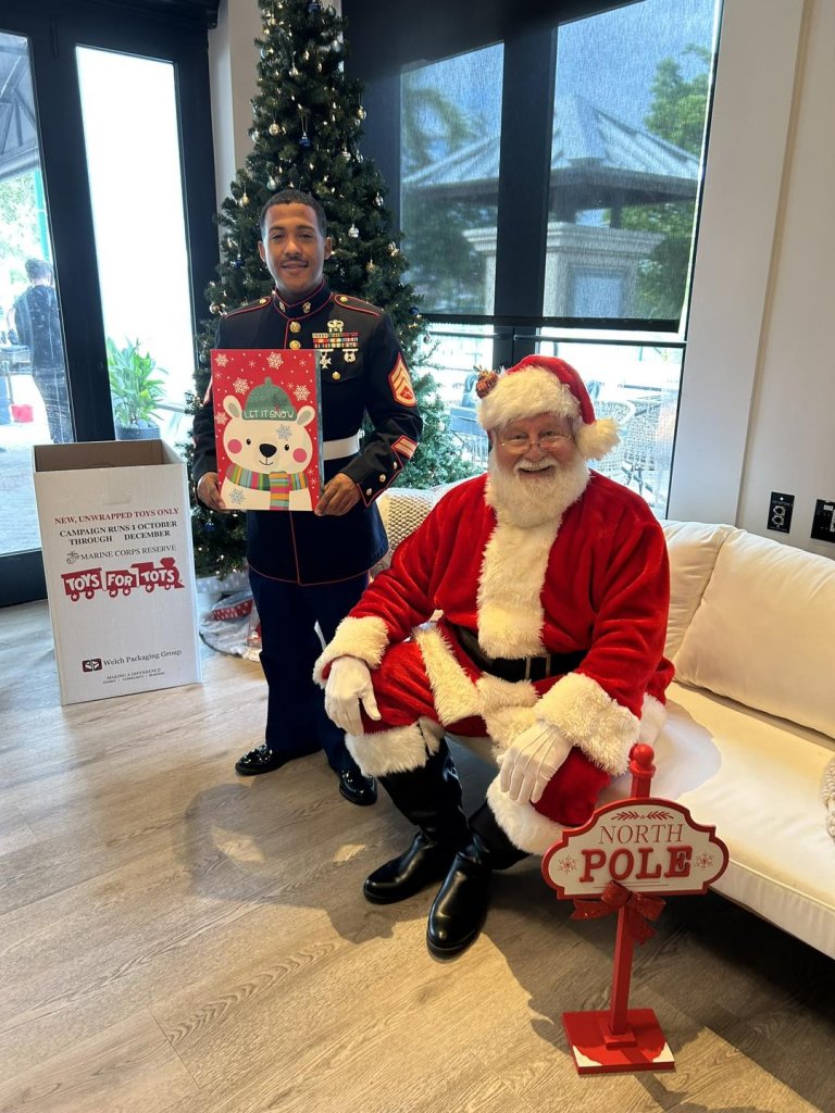 Ein US-Marine steht neben dem Weihnachtsmann während einer Weihnachtsspielzeugaktion mit einer festlichen Geschenktüte in der Hand, mit einem geschmückten Weihnachtsbaum und einer "Toys for Tots"-Spendenbox im Hintergrund