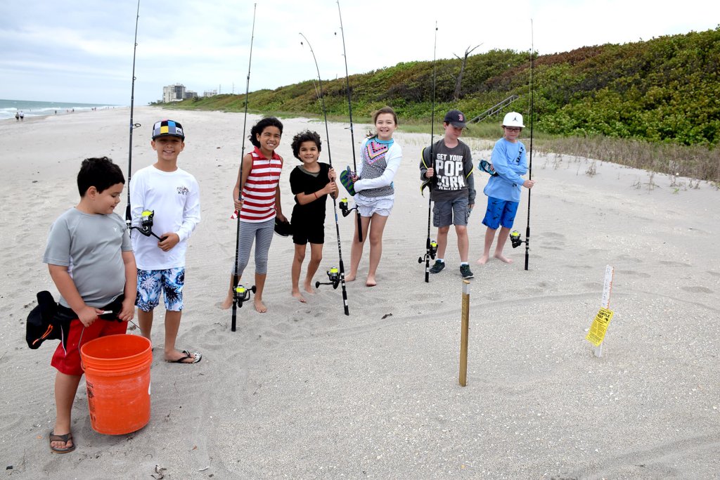 Un grupo de seis niños, todos con cañas de pescar en la mano, posan en una playa de arena con las dunas verdes y el océano como telón de fondo