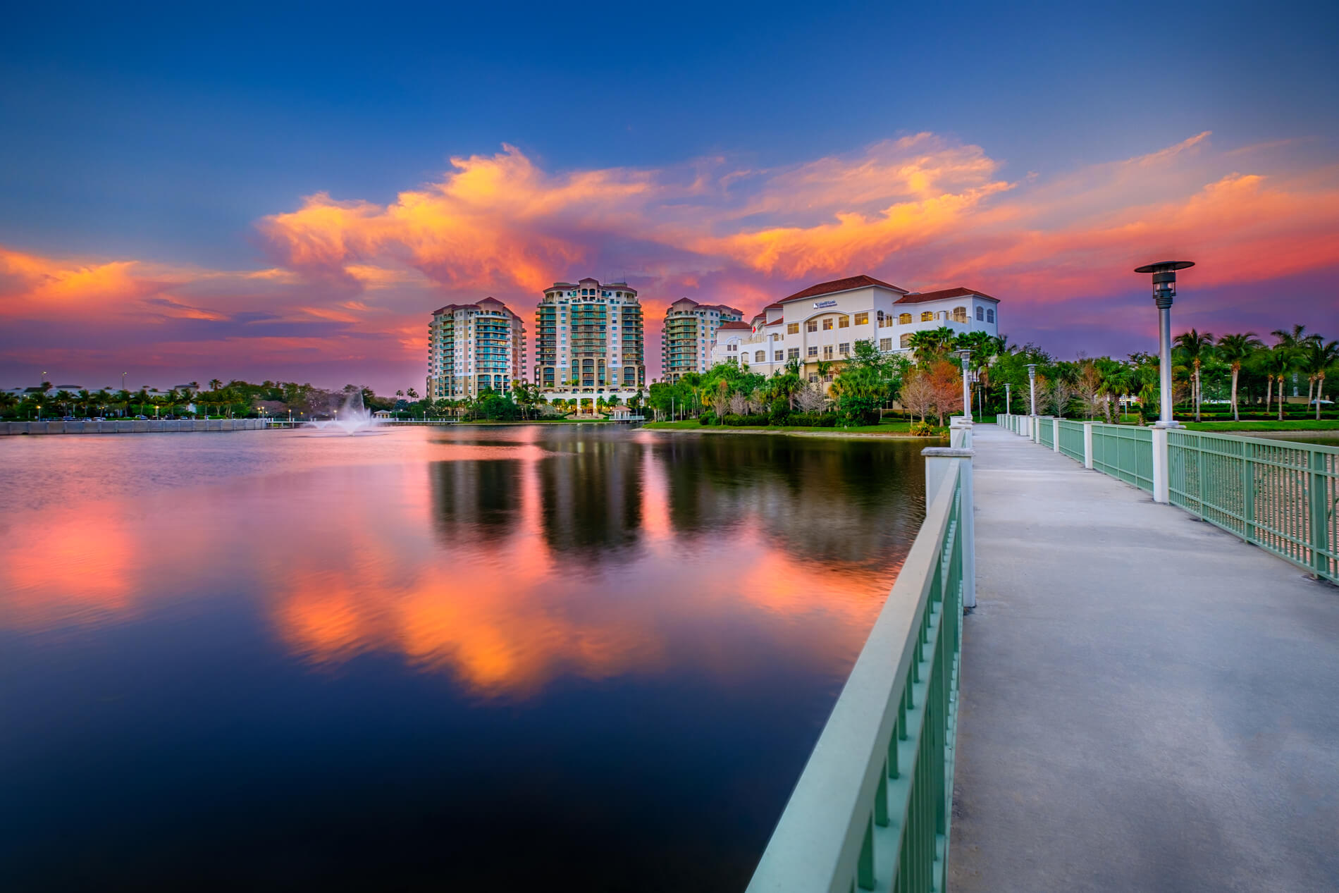 A serene lake reflects the vibrant sunset sky, with modern high-rises and a picturesque white building in the background, all framed by a tranquil walkway