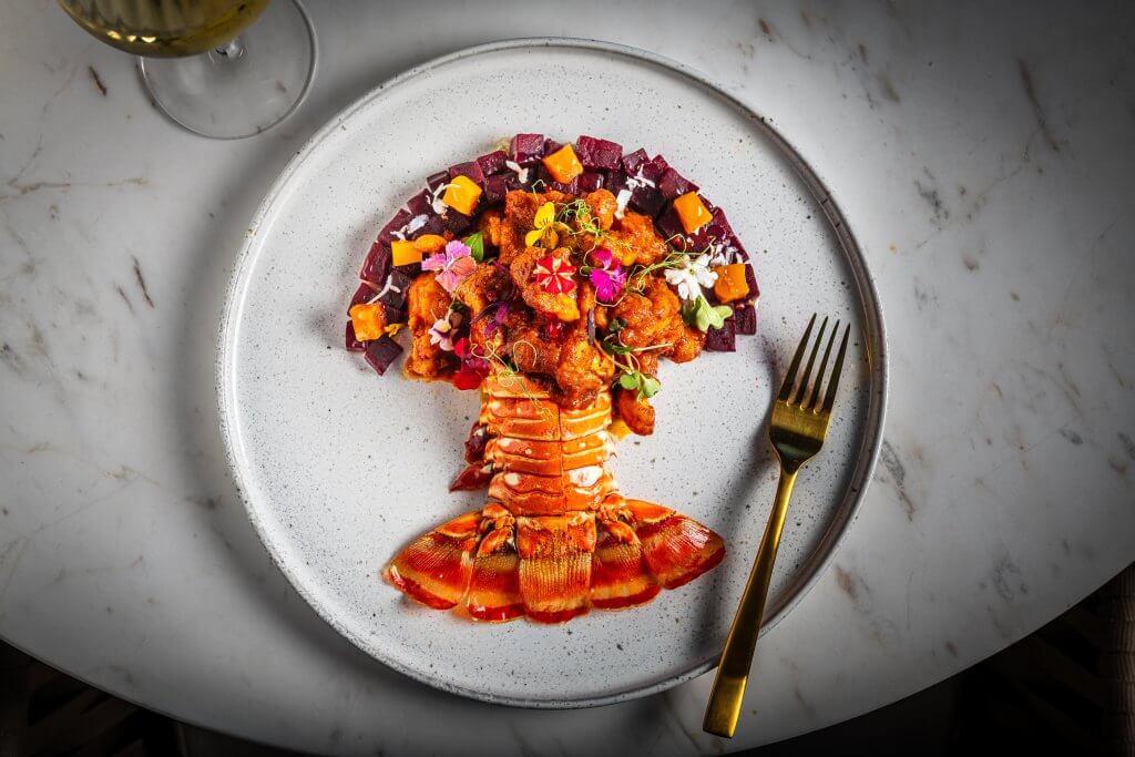 Un plato con una presentación exquisita que incluye una cola de langosta entera con intensos tonos rojos y anaranjados, adornada con coloridos microvegetales y flores comestibles, servida en un plato blanco con un tenedor dorado y una copa de vino blanco