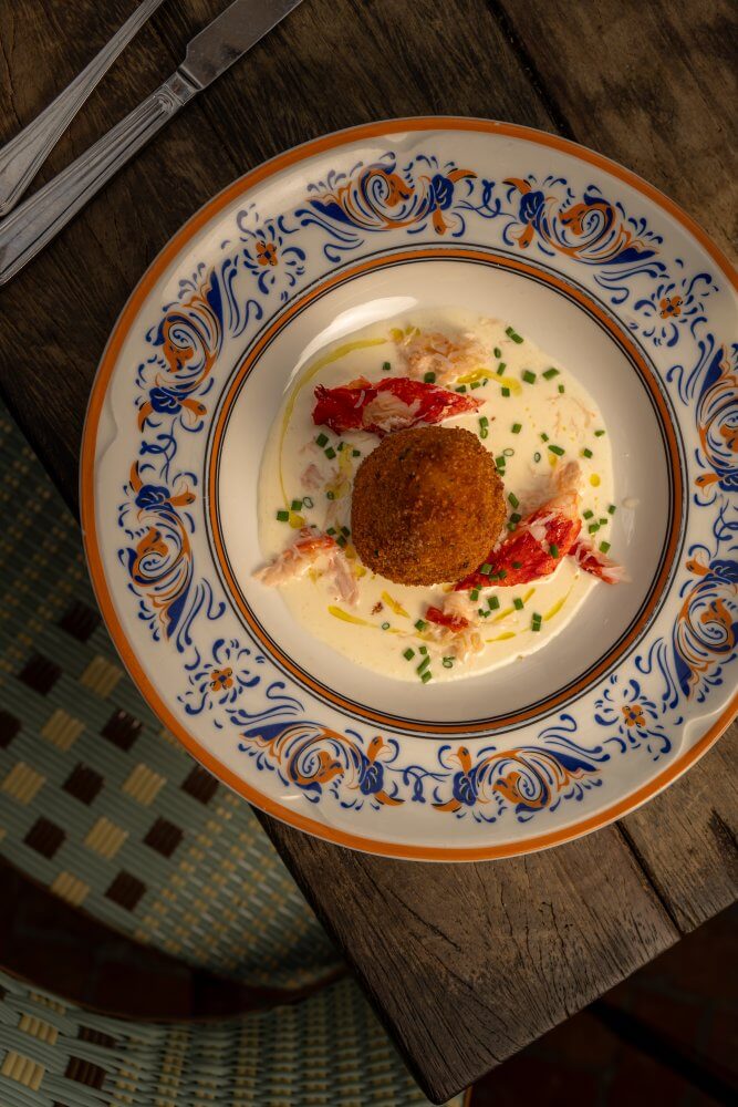 Una bola crujiente de mariscos fritos reposa sobre una salsa cremosa con trozos de langosta y cebollino en un plato decorativo