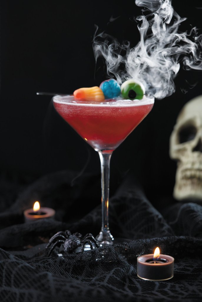 A spooky Halloween cocktail with candy eyeballs and smoke, served in a martini glass on a black lace tablecloth with candles and a skull