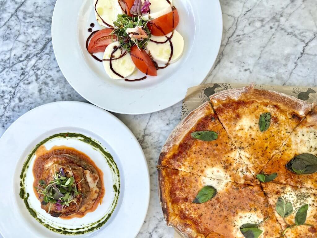 Three Italian dishes: Caprese salad with mozzarella, tomatoes, and balsamic glaze; eggplant parmesan with pesto drizzle; and a margherita pizza with fresh basil