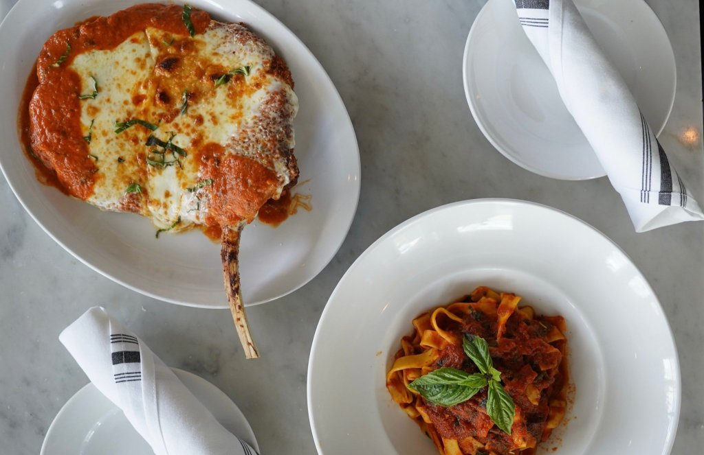 Una mesa con tres platos blancos: uno con una gran porción redonda de lasaña con una corteza crujiente y dorada y queso derretido; otro con una ración de pasta en una sabrosa salsa de tomate adornada con albahaca fresca; y un tercer plato vacío. Junto a los platos hay dos servilletas blancas con rayas negras, cuidadosamente dobladas.