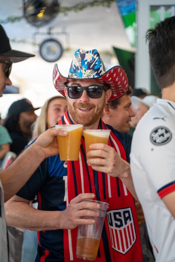 Ein Mann mit patriotischem Cowboyhut und Sonnenbrille hebt zwei Becher Bier in die Höhe und feiert mit einer Menschenmenge bei einer Sportveranstaltung