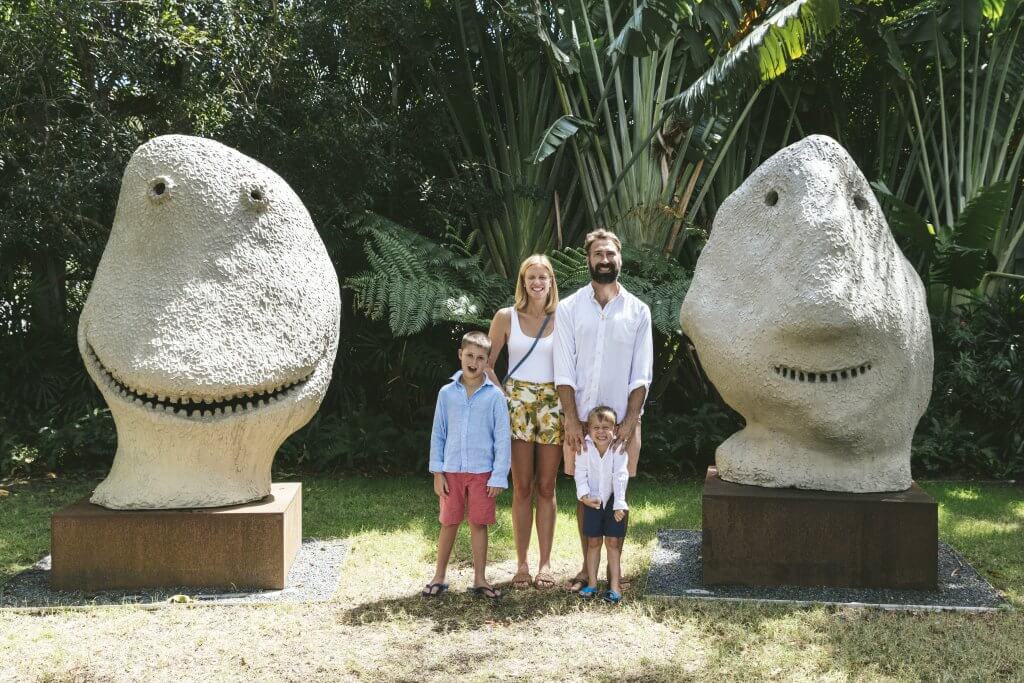 A family of four stands between two large, whimsical stone sculptures shaped like smiling faces in a lush, tropical garden