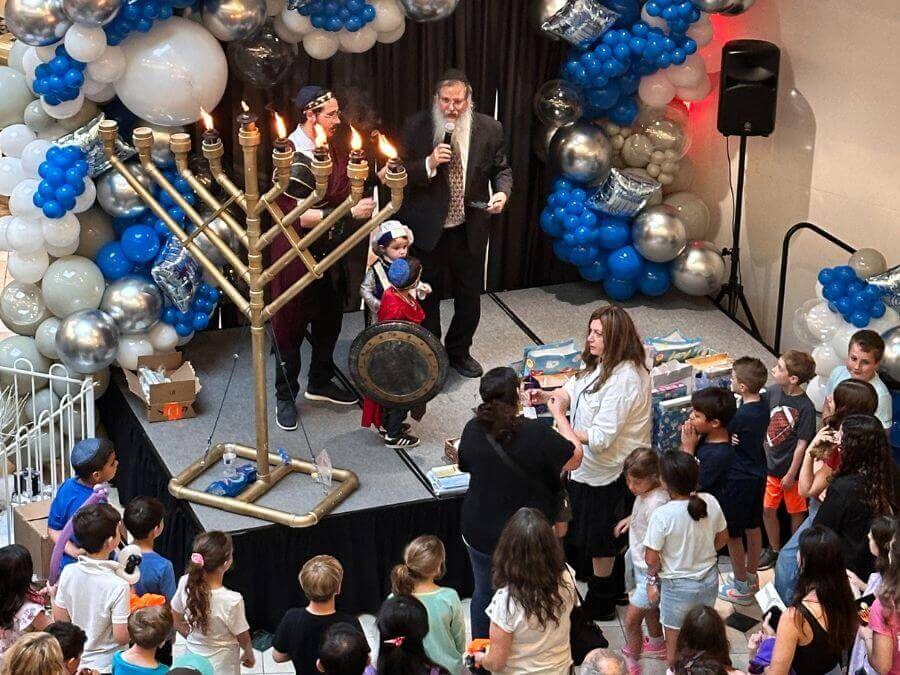 A festive Hanukkah celebration with a large menorah, children dressed in costumes, and a crowd gathered around a stage decorated with balloons