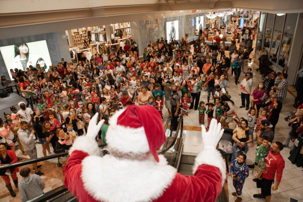 Santa Claus waves to a cheering crowd of children and families from the top of an escalator in a bustling mall during the holiday season