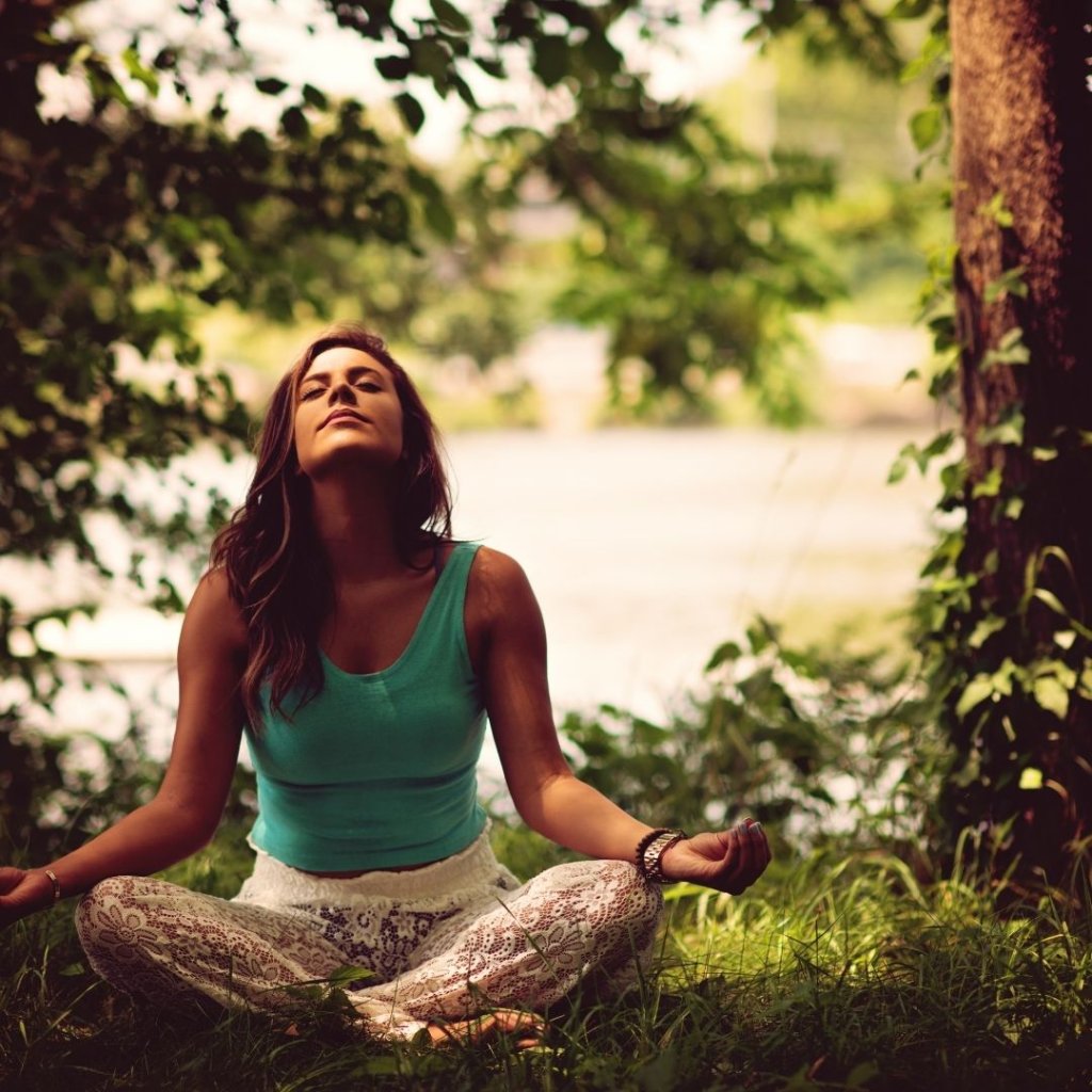 A woman sits cross-legged in a serene outdoor setting, surrounded by lush greenery and a tranquil body of water, practicing meditation with her eyes closed and hands resting on her knees