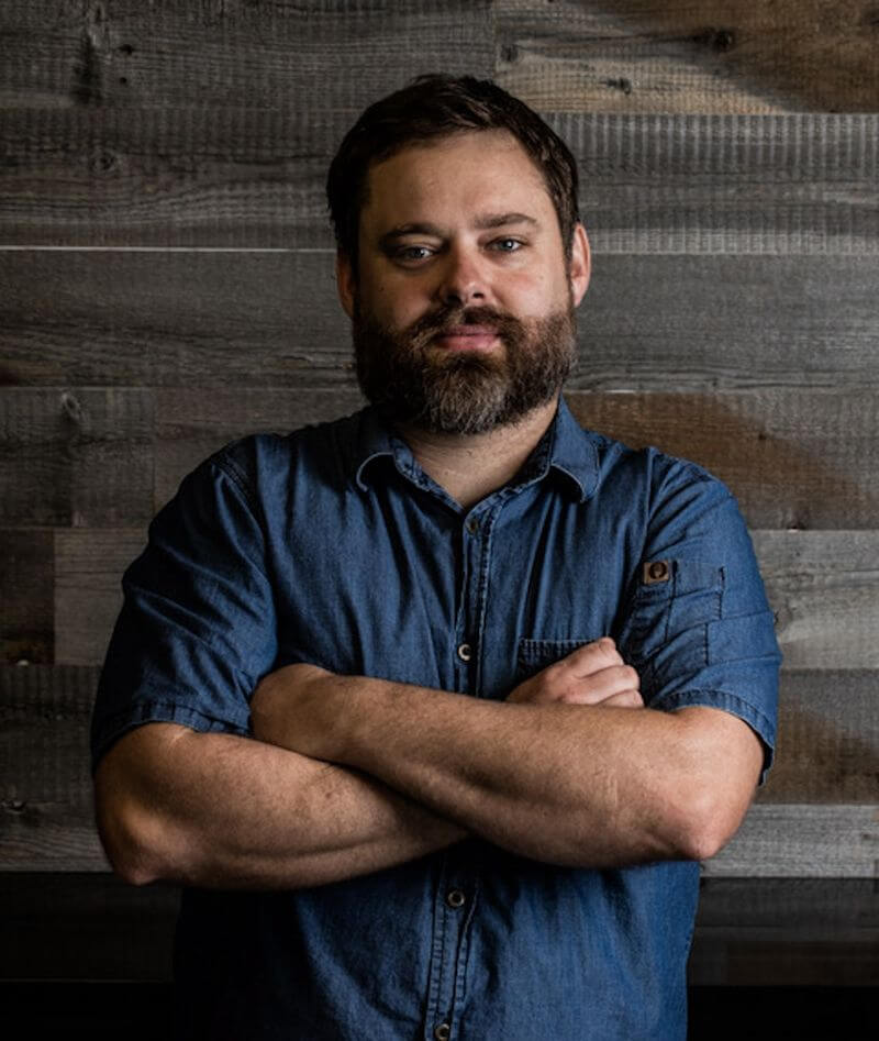 A man with a beard and mustache stands confidently with his arms crossed against a rustic wooden wall