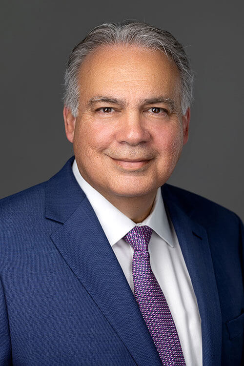 A professional portrait of a man in a blue suit with a purple tie, smiling warmly at the camera
